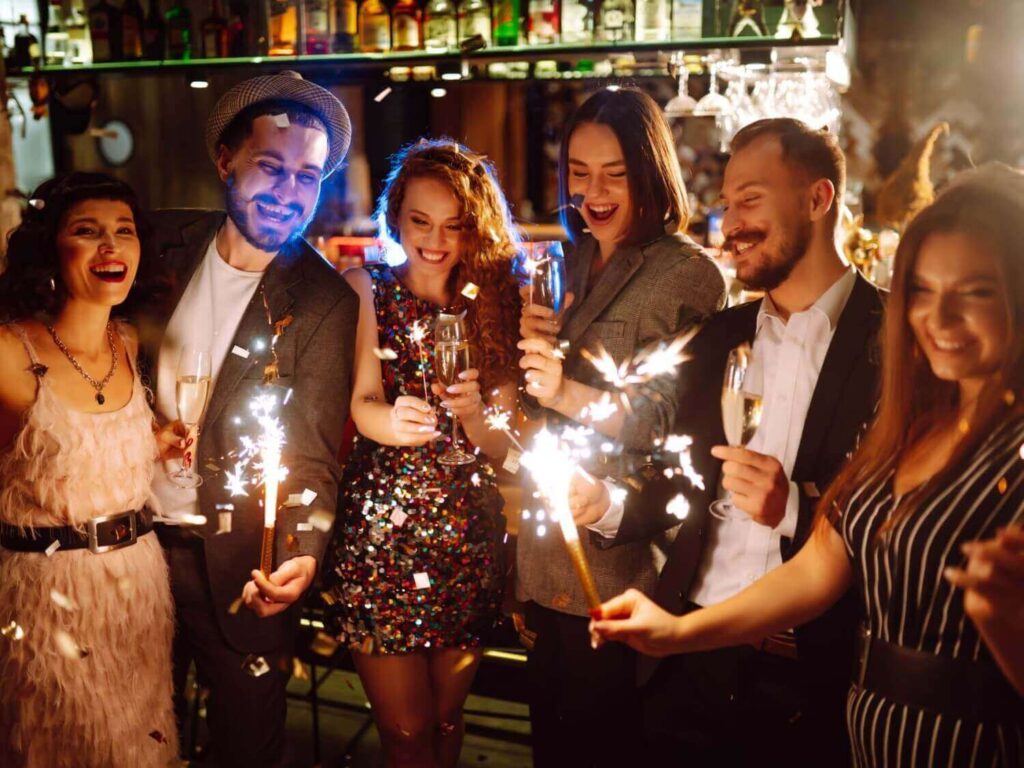 Eine Gruppe von sechs festlich gekleideten Personen lächelt, hält Wunderkerzen und Sektgläser in der Hand und feiert gemeinsam in einer lebhaften, warm beleuchteten Bar während einer Weihnachtsfeier in Wien.