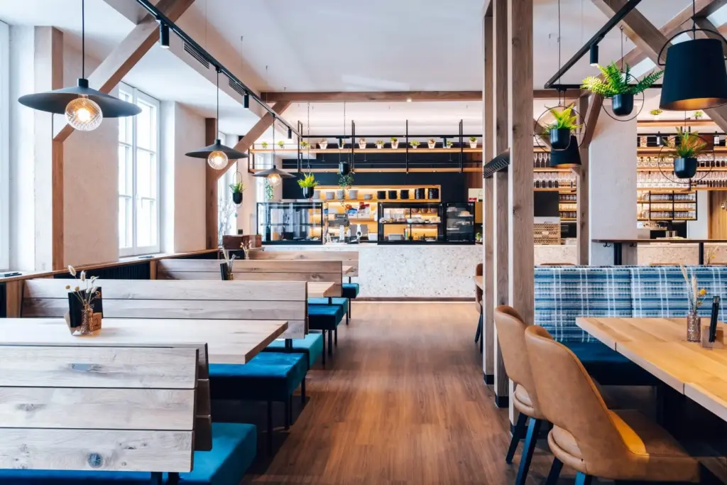 Modernes Restaurantinterieur mit Holztischen und blauen gepolsterten Stühlen auf der linken Seite, braunen Lederstühlen auf der rechten Seite, Hängelampen, Topfpflanzen und einem Tresen mit Regalen und Menütafeln im Hintergrund.