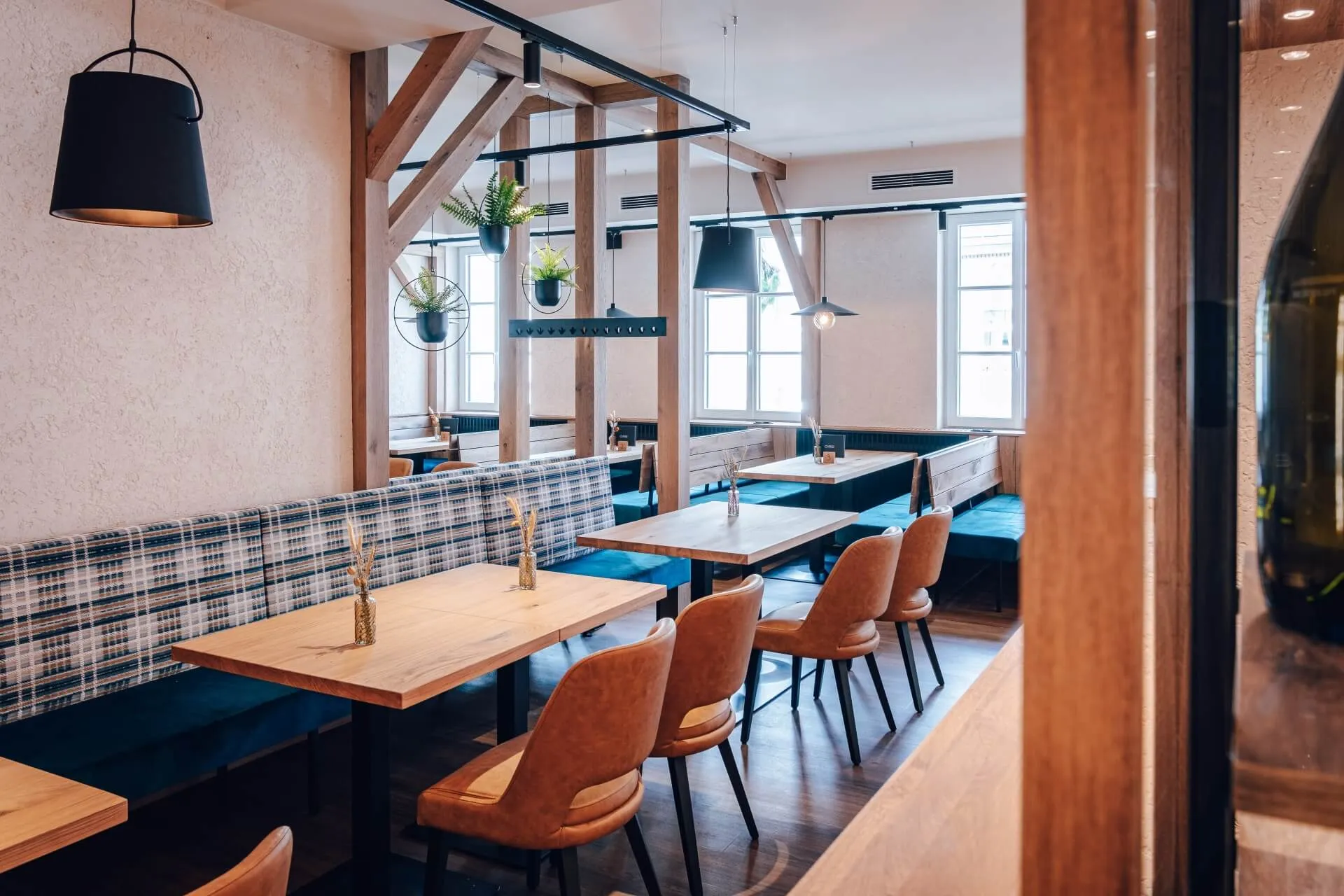 Ein modernes, gemütliches Restaurant mit Holztischen, braunen gepolsterten Stühlen, karierten Sitzbänken, großen Fenstern für natürliches Licht und schwarzen Hängelampen an der Decke.