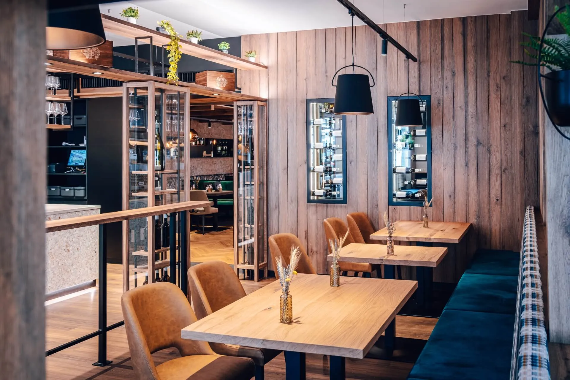 Modernes Restaurant mit Holzwänden, hellen Holztischen, braunen Stühlen, blau gepolsterten Sitzbänken, schwarzen Hängelampen und dekorativen Pflanzen in Regalen. Die Atmosphäre ist sauber und einladend.