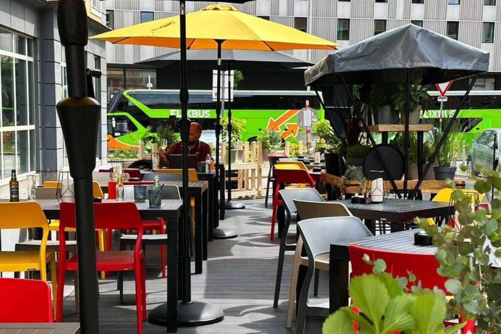 Außencafé Schanigarten mit bunten Stühlen und Tischen, großen gelben Sonnenschirmen und üppigem Grün. Eine Person sitzt an einem Tisch, während ein hellgrüner FlixBus in der Nähe eines modernen Gebäudes im Hintergrund geparkt ist.