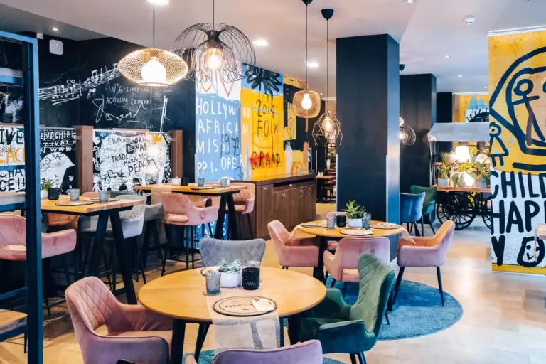 Die moderne Einrichtung des Cafés mit runden Holztischen, bunten gepolsterten Stühlen, Hängelampen und lebendiger Graffiti-Kunst an den Wänden schafft eine lebendige und eklektische Atmosphäre.
