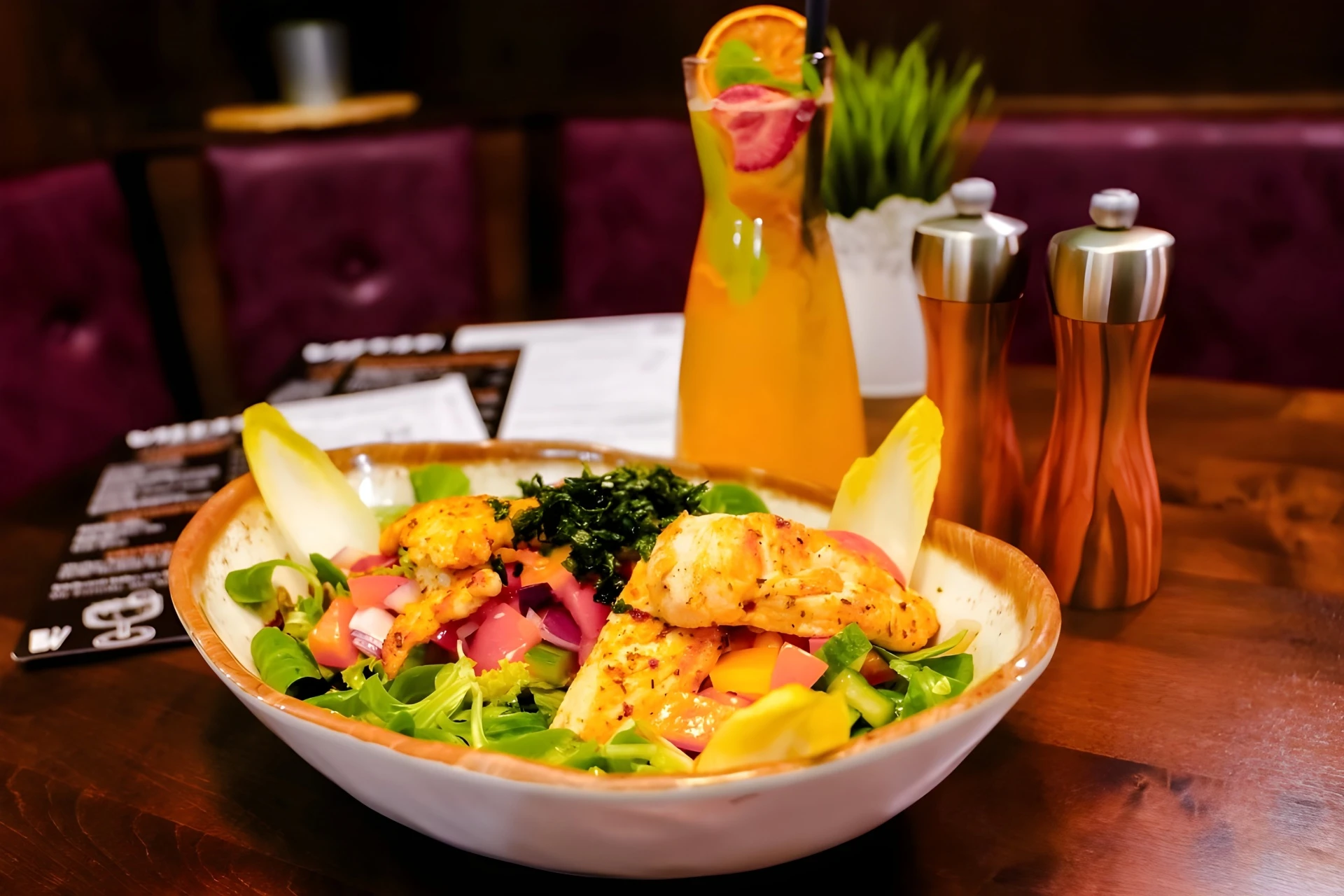 Eine Schüssel mit gegrilltem Hähnchensalat mit gemischtem Grün, Endivien, roten Zwiebeln und Beilagen steht auf einem Holztisch, daneben ein hohes Glas Orangengetränk, Salz- und Pfefferstreuer und eine kleine Pflanze im Hintergrund.