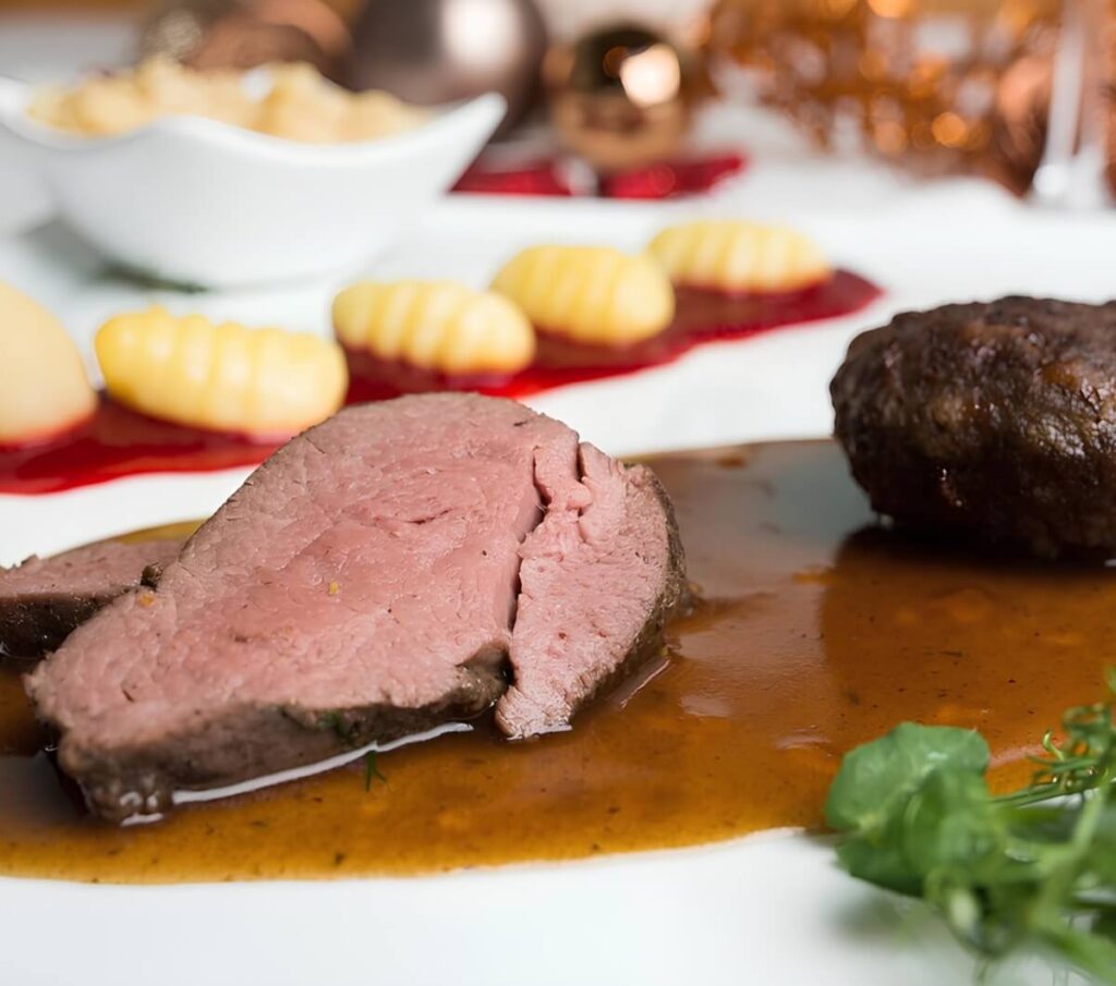 Eine Nahaufnahme eines aufgeschnittenen Bratens mit brauner Soße auf einem weißen Teller, serviert mit Gnocchi und einer Beilage im Hintergrund, garniert mit Grünzeug - ein echter Wiener Restaurant Geheimtipp.