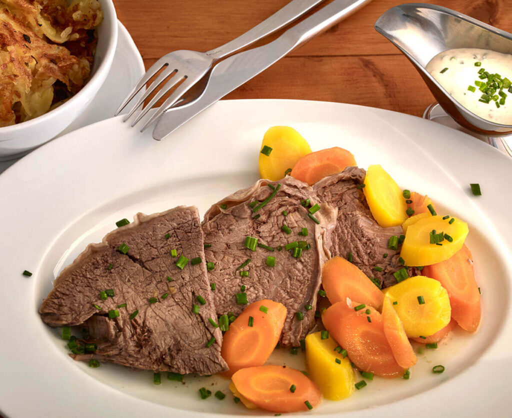 Gekochtes Rindfleisch in Scheiben, serviert mit gelben und orangefarbenen Karotten, garniert mit Schnittlauch auf einem weißen Teller - entdecken Sie dieses klassische Gericht in einem Wiener Restaurant Geheimtipp. Gabel und Messer liegen über dem Teller, daneben stehen Soße und eine Schüssel mit Kartoffeln.
