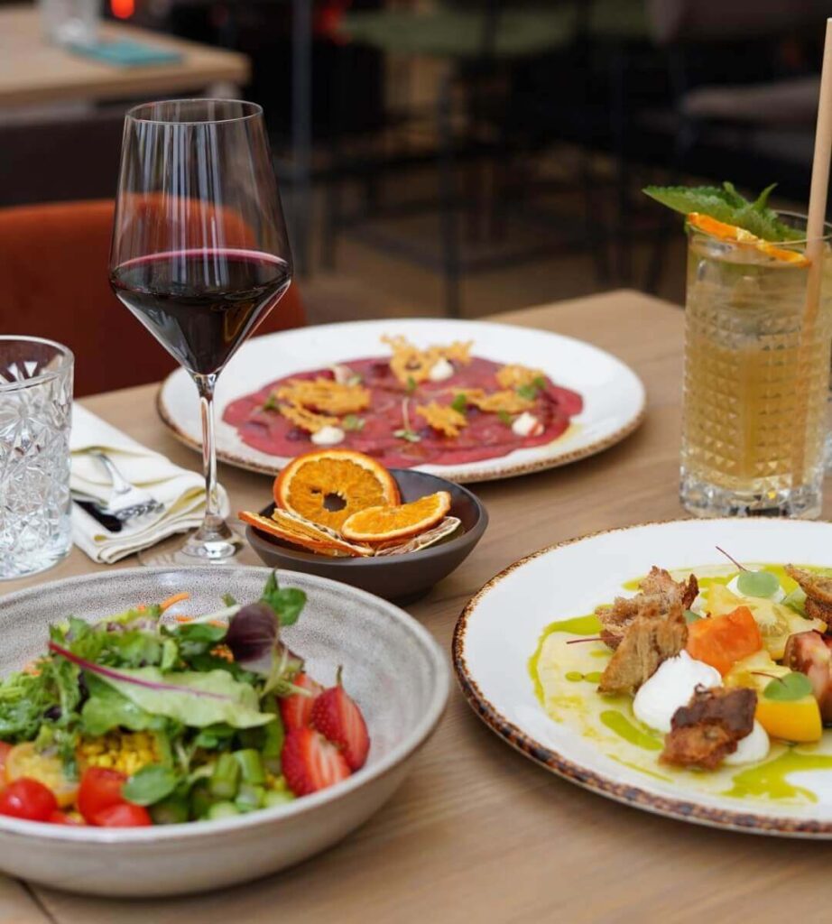 Auf einem gedeckten Tisch in einem Wiener Restaurant Geheimtipp stehen ein Glas Rotwein, ein Cocktail, Salat mit Erdbeeren, dünn geschnittenes Fleisch mit Beilage, gebratenes Fleisch mit Sauce und Gemüse sowie getrocknete Orangenscheiben in einer kleinen Schale.