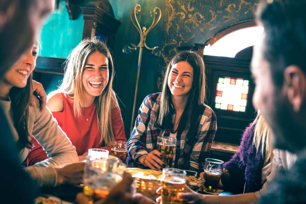 Eine Gruppe von Freunden sitzt um einen Tisch in einem Pub, lacht und trinkt gemeinsam. Teller mit Snacks und Gläser mit Bier füllen den Tisch und schaffen eine lebhafte Atmosphäre - perfekt für diejenigen, die partyraum mieten Wien suchen.