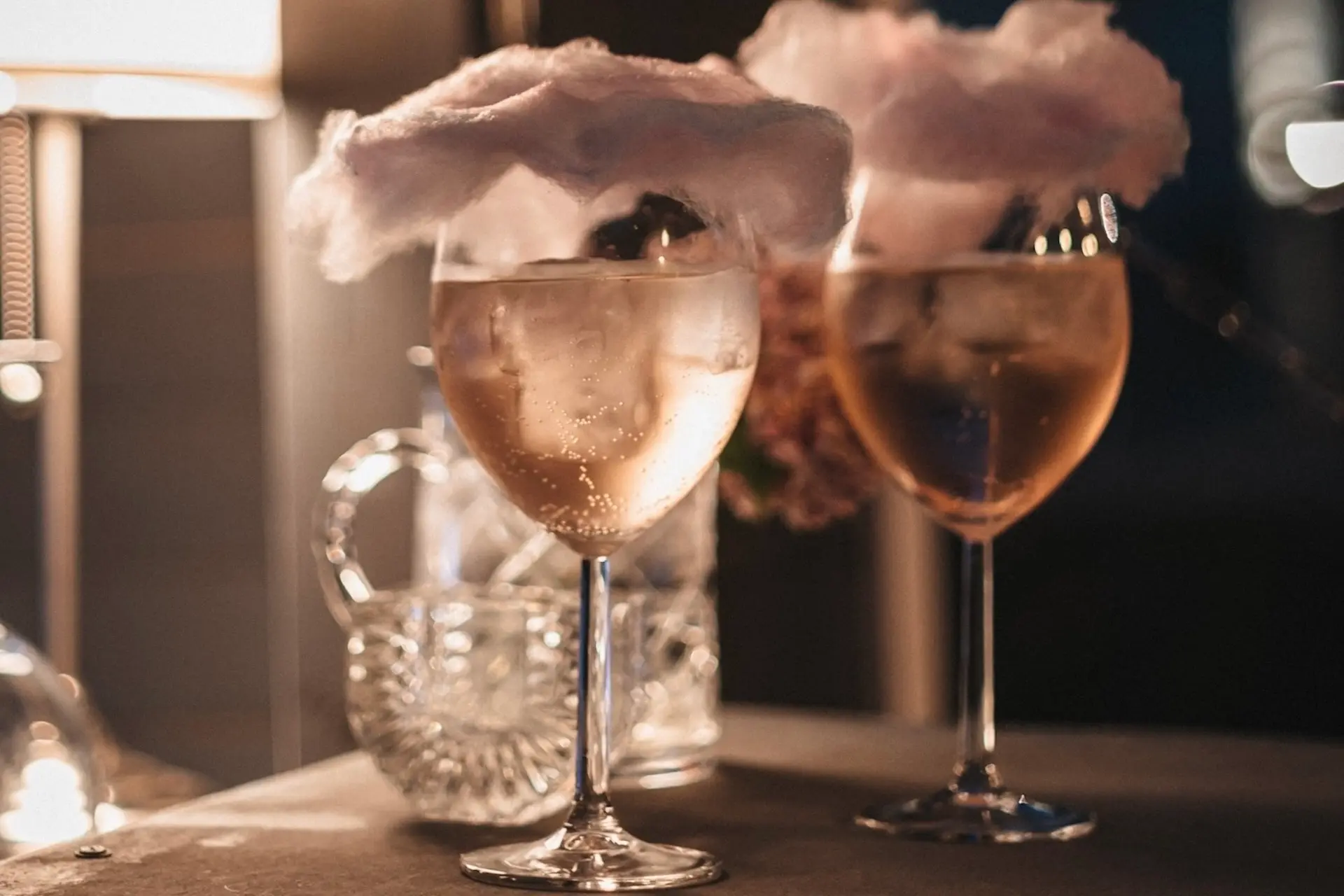 Zwei Weingläser, gefüllt mit einem hellen Getränk, sind mit flauschiger Zuckerwatte bestückt. Ein Glaskrug und eine weiche, warme Beleuchtung im Hintergrund schaffen eine gemütliche und elegante Atmosphäre.