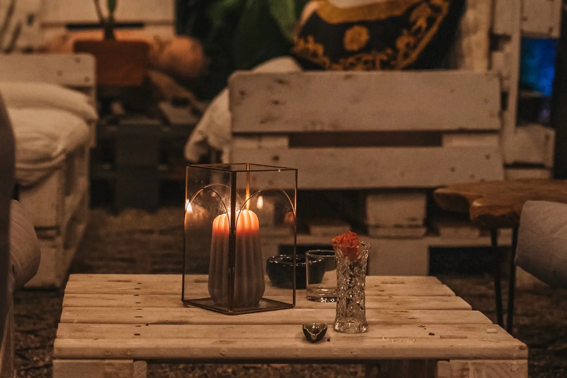 Ein gemütlicher Raum mit einem Holztisch mit einer brennenden Kerze in einer Glaslaterne, einer kleinen Vase mit einer roten Blume und einem Getränk, umgeben von weißen Palettenmöbeln.