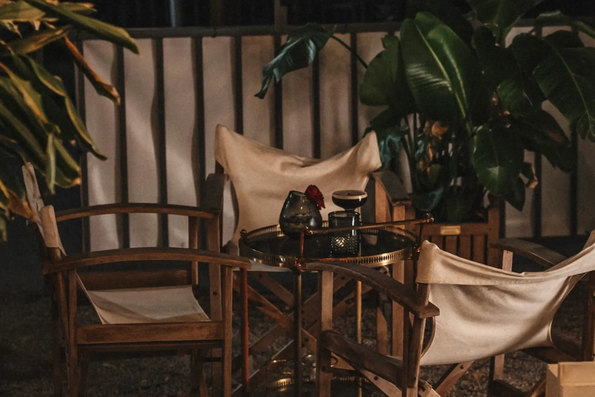 Ein gemütlicher nächtlicher Sitzplatz im Freien mit Holzstühlen, einem runden Tisch mit einer Kerze und einer Blumenvase, umgeben von üppigen Grünpflanzen und sanfter Beleuchtung.