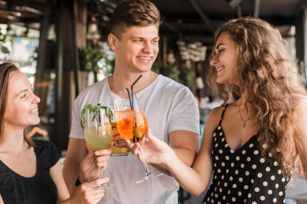 Drei junge Erwachsene lächeln und stoßen mit bunten Cocktails in einer lebhaften, modernen Innenumgebung an. Inspiriert von lustigen Cocktail-Rezepten, trägt eine Frau ein schwarzes gepunktetes Kleid, während alle die lebhafte, fröhliche Atmosphäre genießen.