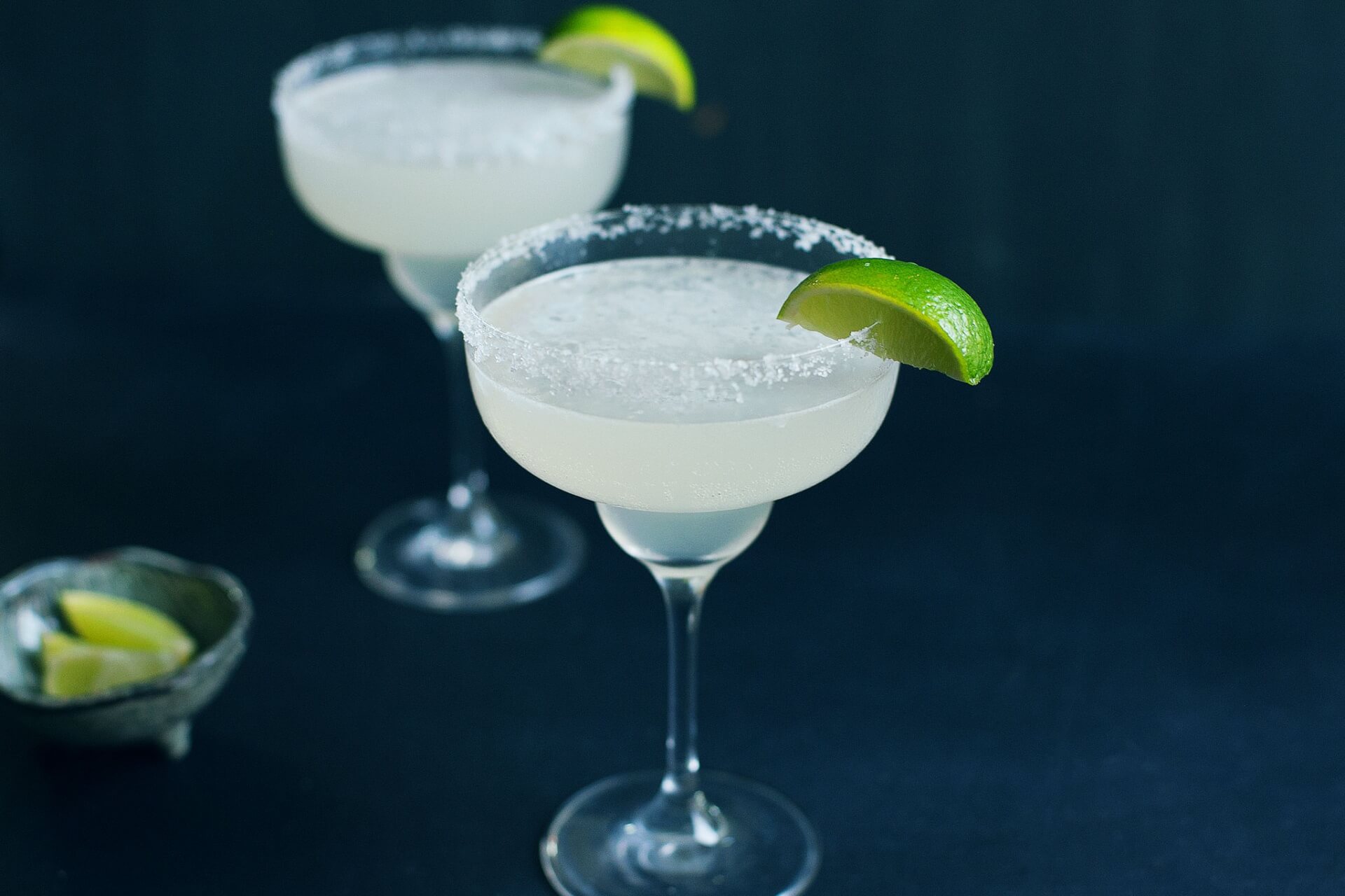 Zwei Margarita-Cocktails in salzgerahmten Gläsern, jeweils mit einer Limettenspalte garniert, stehen auf einer dunklen Fläche. Im Hintergrund verschwimmt eine kleine Schale mit Limettenspalten - die perfekte Inspiration für Ihr nächstes Cocktail-Rezepte-Abenteuer.