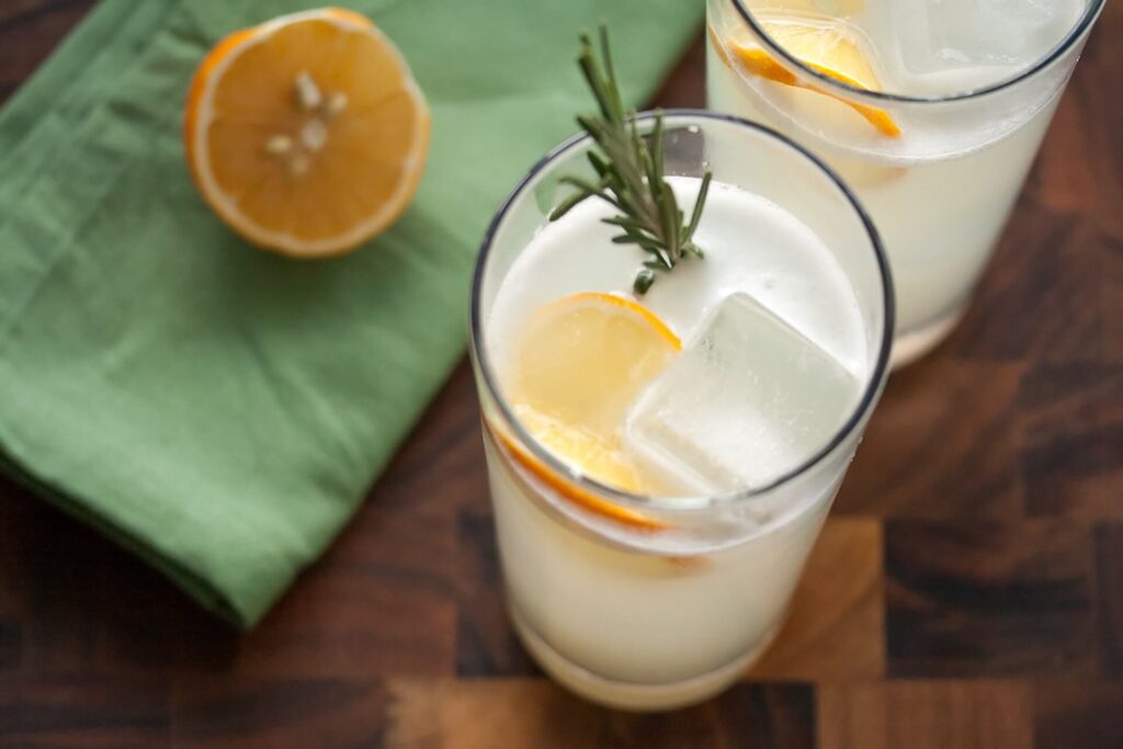 Zwei Gläser eines hellgelben Drinks mit Eiswürfeln, Zitronenscheiben und einem Zweig Rosmarin. Im Hintergrund liegt eine halbierte Zitrone auf einem grünen Tuch - die perfekte Inspiration für Ihre nächsten Cocktail-Rezepte.