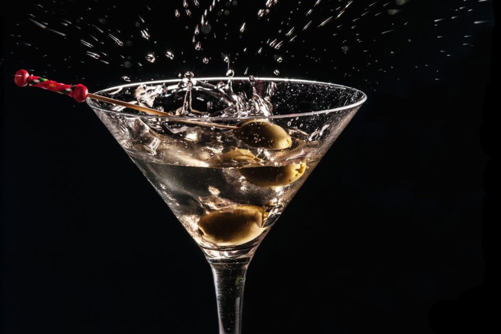 Ein Spritzer fängt sich in der Luft, als Oliven in ein mit klarer Flüssigkeit gefülltes Martiniglas fallen, garniert mit einem roten Cocktail-Pick - ein eleganter Moment, der sich perfekt für jede Cocktail-Rezepte-Kollektion eignet, vor einem schlichten schwarzen Hintergrund.