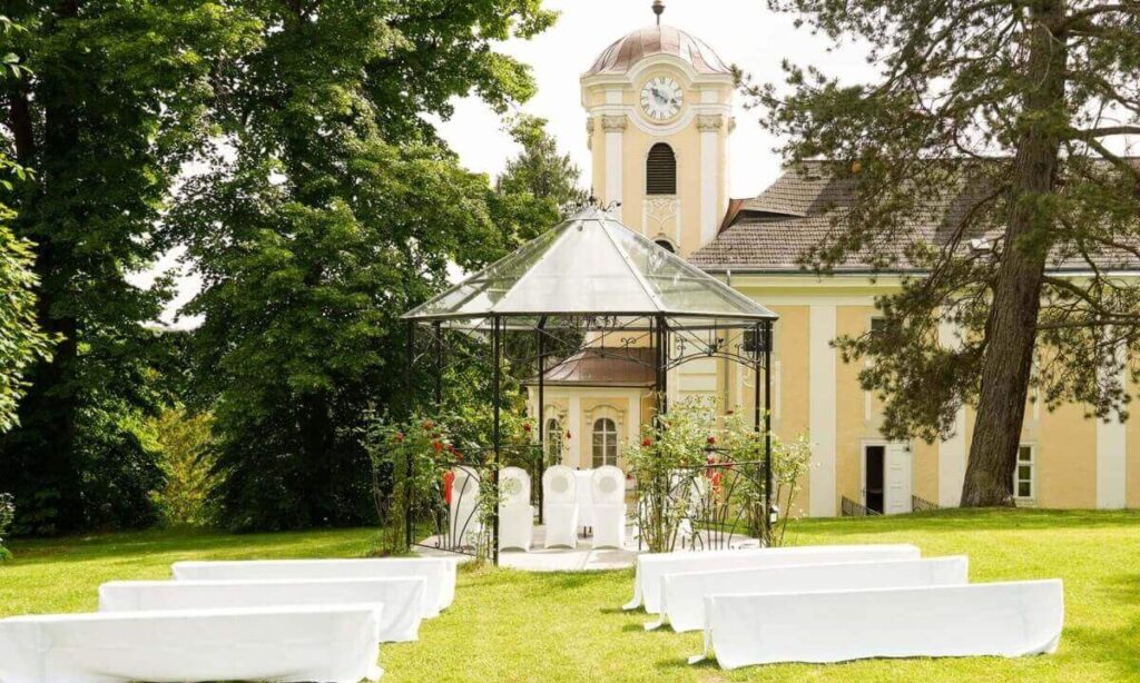 Eine Hochzeitszeremonie auf einer Wiese in einer bezaubernden Hochzeitslocation in Niederösterreich mit weißen Bänken vor einem Glaspavillon mit zwei weißen Stühlen im Inneren, vor einem gelben Gebäude mit einem Uhrenturm und umgeben von grünen Bäumen.