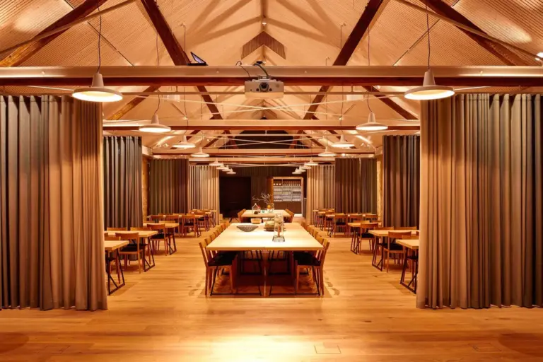 Warm beleuchteter Speisesaal mit Holzböden, hoher Gewölbedecke, sichtbaren Balken, mehreren langen Tischen mit Stühlen, Pendelleuchten und raumhohen Vorhängen an den Seiten, die eine gemütliche, einladende Atmosphäre schaffen.