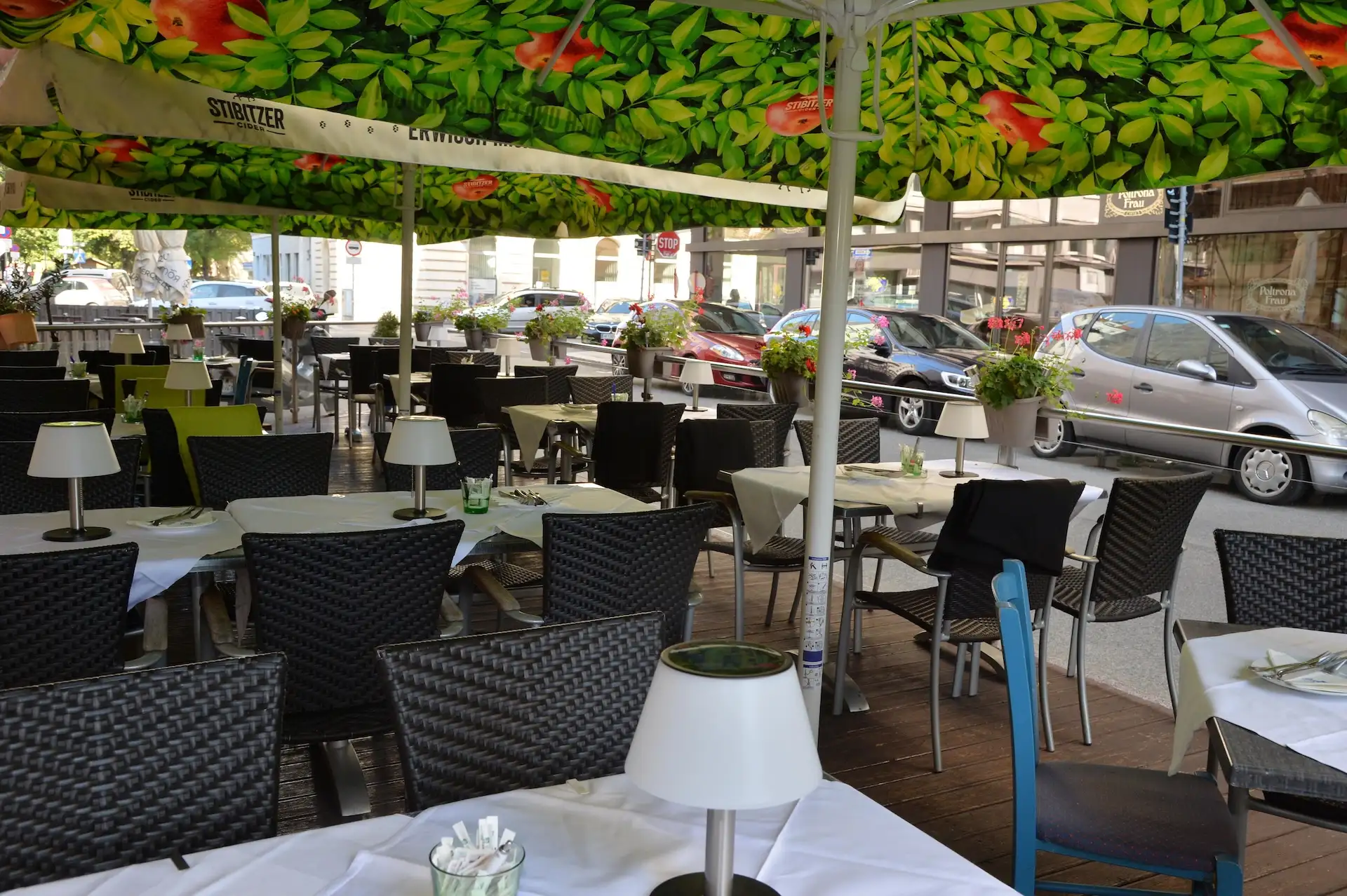 Café im Freien mit schwarzen Korbstühlen und Tischen mit weißem Tischtuch unter einem grünen Vordach. Auf jedem Tisch stehen kleine Lampen. Das Café liegt an einer Stadtstraße mit geparkten Autos und nahe gelegenen Gebäuden.