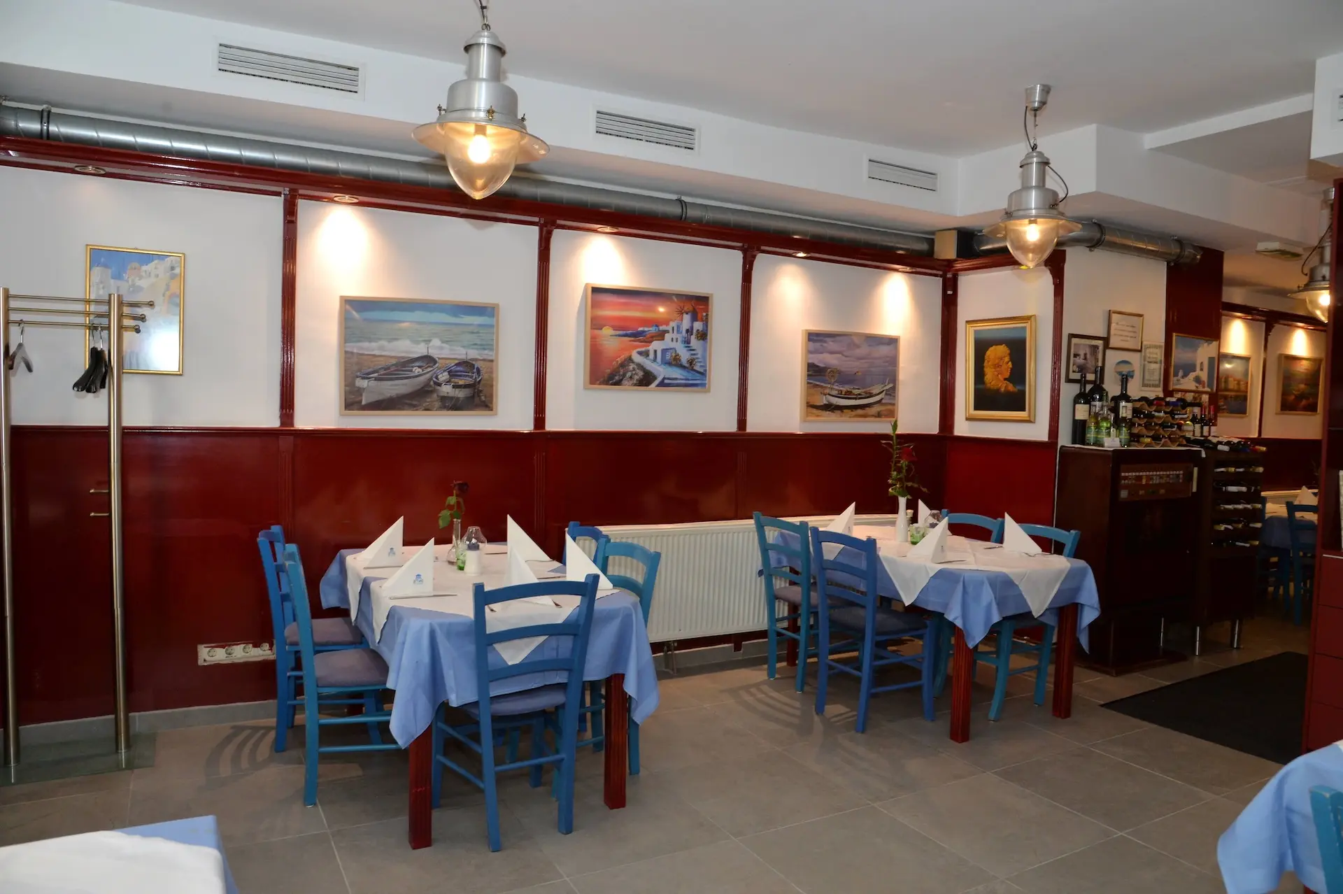 Ein gemütliches Restaurant mit blauen Stühlen, blau-weiß gedeckten Tischen, Gemälden an rot getäfelten Wänden, modernen Hängelampen und einem kleinen Barbereich im Hintergrund.