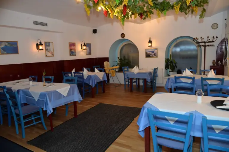 Ein gemütliches Restaurant mit blauen Stühlen und Tischen mit weißen und blauen Tischdecken, dekoriert mit hängenden Weinreben an der Decke und gerahmten Bildern an den Wänden. Der Raum ist sanft beleuchtet und menschenleer.