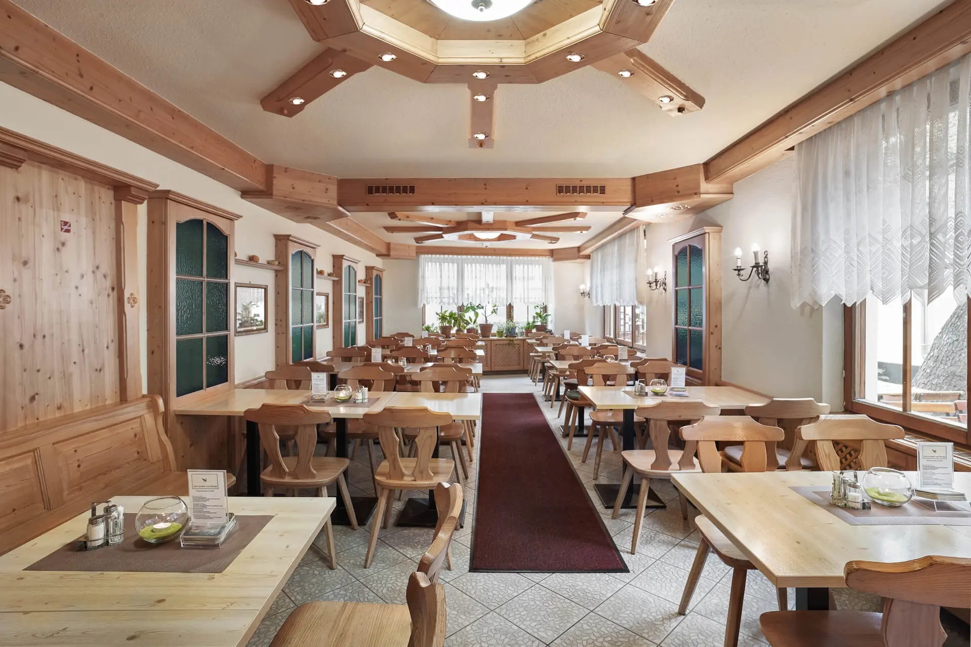 Ein heller, geräumiger Speisesaal mit Holztischen und -stühlen, gefliesten Böden, großen Fenstern mit transparenten Vorhängen und einer dekorativen Holzdecke mit Beleuchtung.