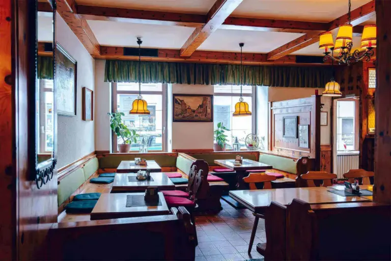 Ein gemütliches, rustikales Restaurant mit Holzbalken, Tischen und Bänken. Das Sonnenlicht strömt durch große Fenster, und darüber hängen gelbe Hängelampen. Rote und grüne Kissen säumen die Sitzbereiche.