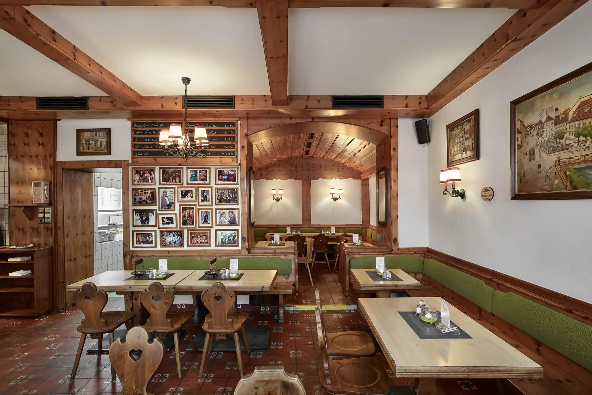 Ein gemütliches Restaurant mit Holzbalken, grün gepolsterten Bänken, Holzstühlen und Tischen. Gerahmte Fotos und Gemälde schmücken die Wände, und die sanfte Beleuchtung schafft eine warme, einladende Atmosphäre.