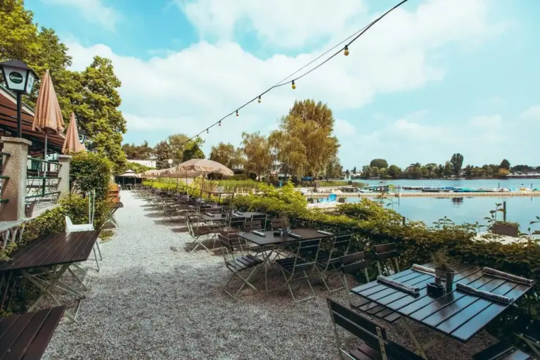 Ein Café im Freien am See mit leeren Tischen und Stühlen auf einem Kiesweg, Lichterketten über dem Kopf, üppigem Grün und Menschen, die im See schwimmen, unter einem blauen Himmel mit vereinzelten Wolken.