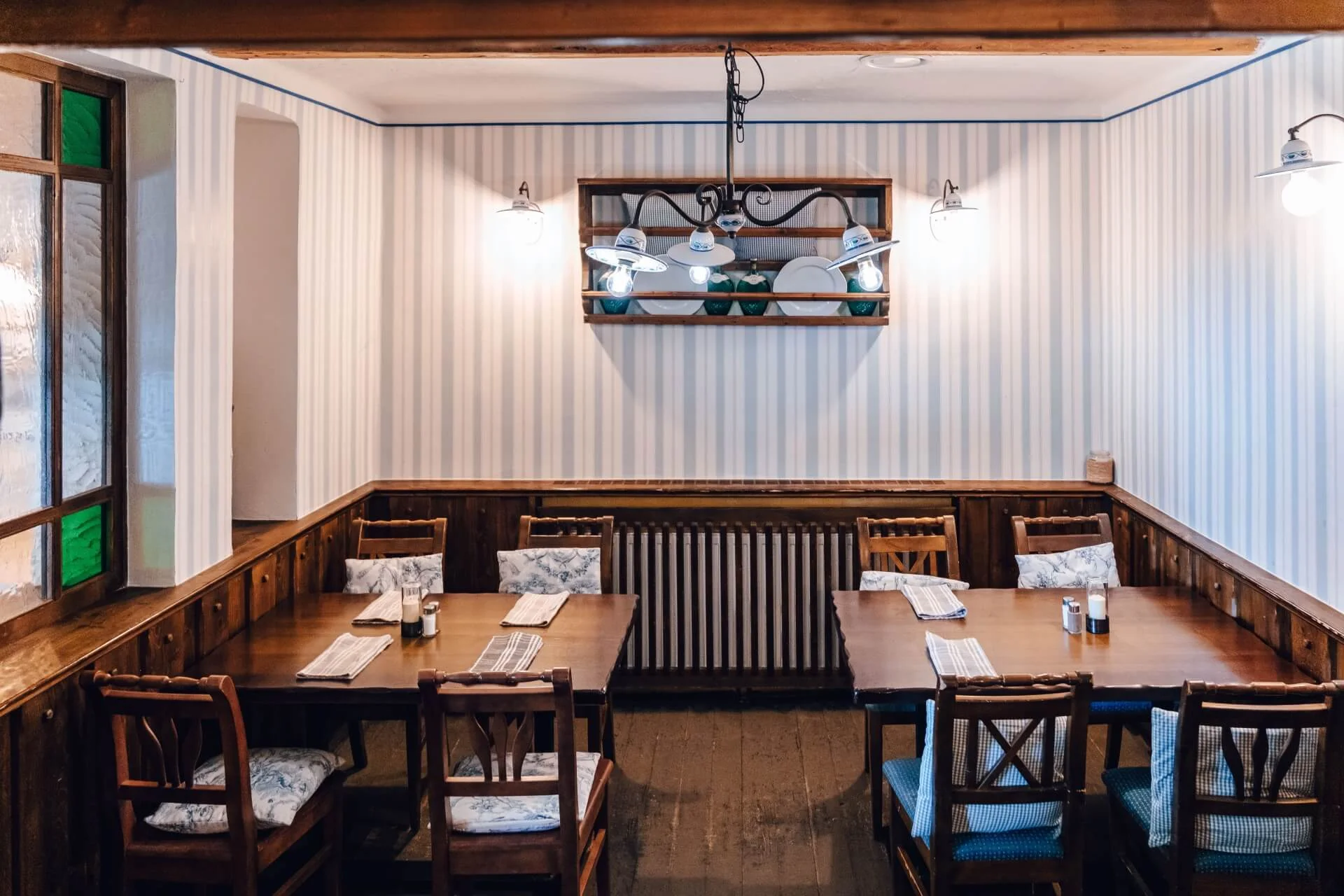 Ein gemütlicher Restaurantbereich mit Holztischen und -stühlen, gestreifter Tapete, Kissen auf den Sitzen und dekorativen Tellern auf einem Regal über einem Heizkörper. Sanfte Beleuchtung schafft eine warme, einladende Atmosphäre.