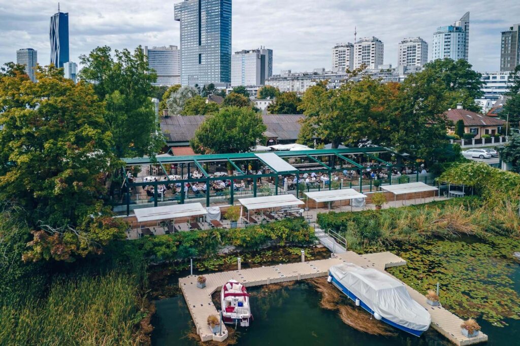 Ein Restaurant am Flussufer mit Sitzgelegenheiten im Freien ist von Bäumen umgeben, und an einer kleinen Anlegestelle davor liegen Boote vor Anker. Im Hintergrund sind hohe moderne Gebäude und die Skyline der Stadt bei bewölktem Himmel zu sehen.