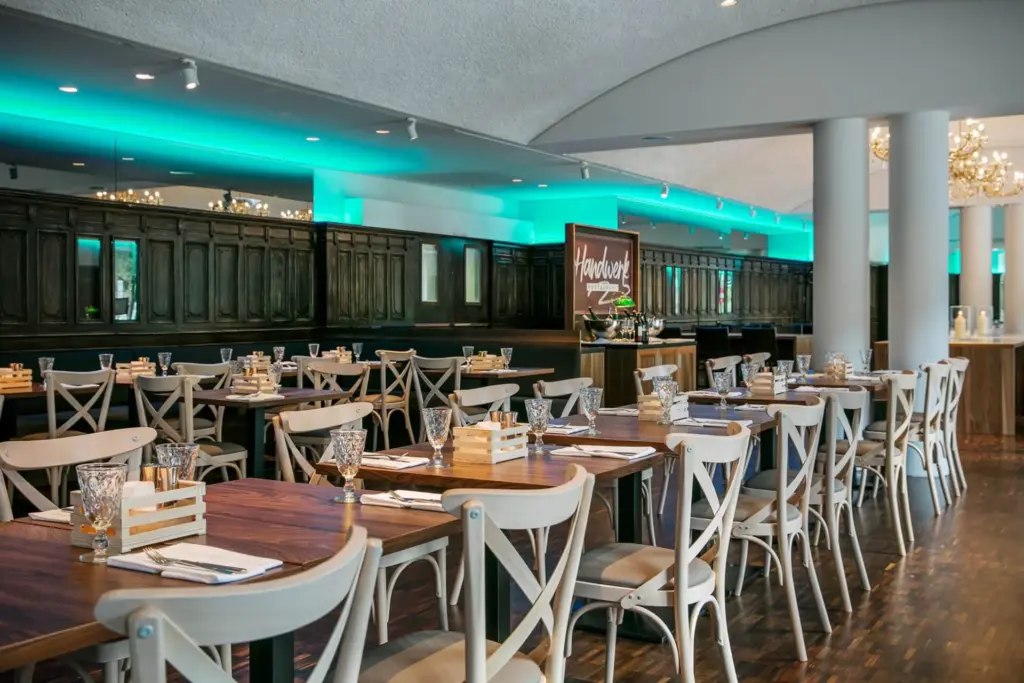 Ein modernes Restaurant mit Holztischen und weißen Stühlen, die mit Geschirr und Gläsern ausgestattet sind. Der Raum ist mit türkisfarbener Beleuchtung, hölzernen Akzenten und einem Schild mit der Aufschrift "Hardtwald" ausgestattet.