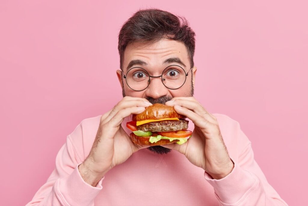 Ein Mann mit Brille und Bart, der ein rosafarbenes Sweatshirt trägt, hält einen großen Hamburger mit Salat, Tomate, Käse und Rinderpastete in der Hand - und genießt einen leckeren Bissen während der Board Night. Der Hintergrund ist einfarbig rosa.