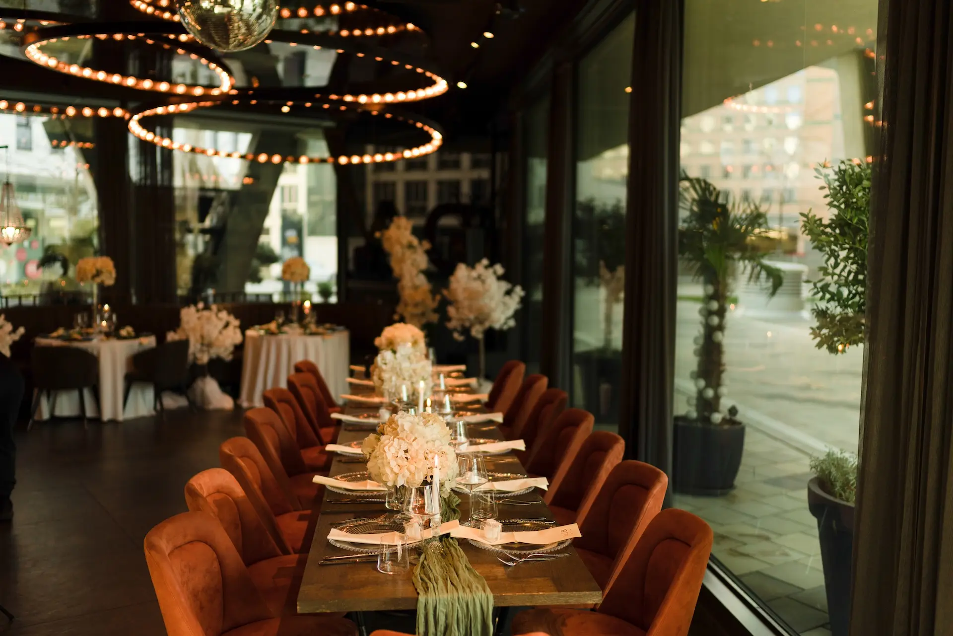 Ein langer Esstisch mit orangefarbenen Samtstühlen ist für eine formelle Veranstaltung gedeckt und mit weißen Blumenkränzen dekoriert. Darüber hängen warme Lichter, und große Fenster geben den Blick auf die Terrasse und die Stadt frei.