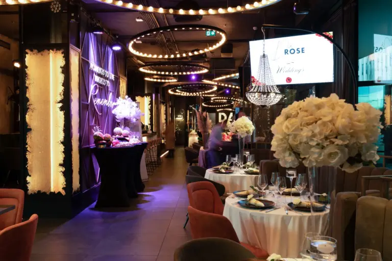 Elegantes Restaurantinterieur, dekoriert für eine Hochzeit mit runden Tischen, weißen Blumenkränzen, Kronleuchtern und Umgebungsbeleuchtung. Eine Person steht in der Nähe eines Desserttisches, und auf einem großen Bildschirm werden die Details der Hochzeitsfeier angezeigt.