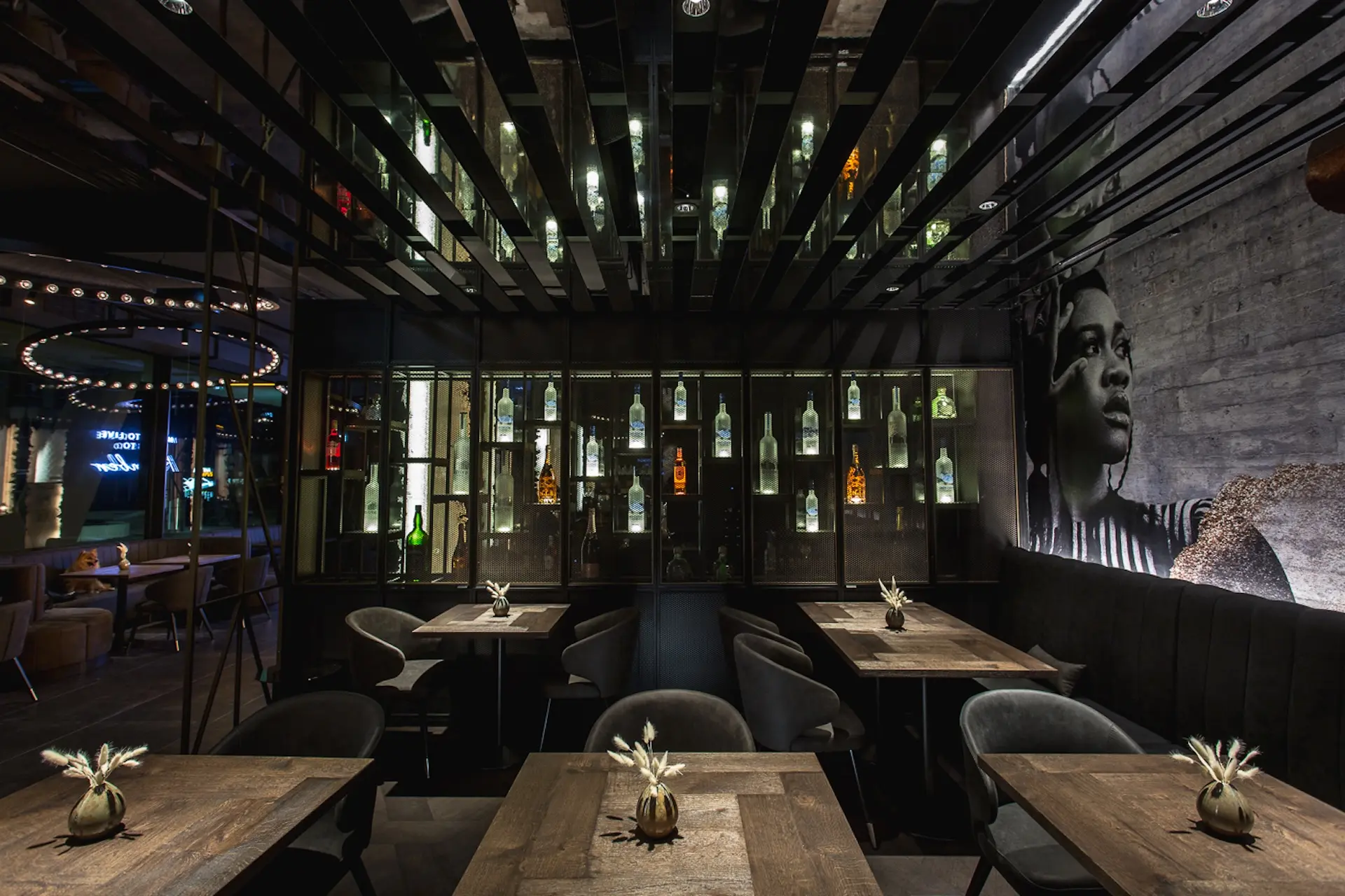 Ein schummriges, modernes Restaurant mit Holztischen, schwarzen Stühlen, dekorativen Flaschen in Regalen und einem großen schwarz-weißen Wandgemälde mit einem Frauengesicht an der Wand.