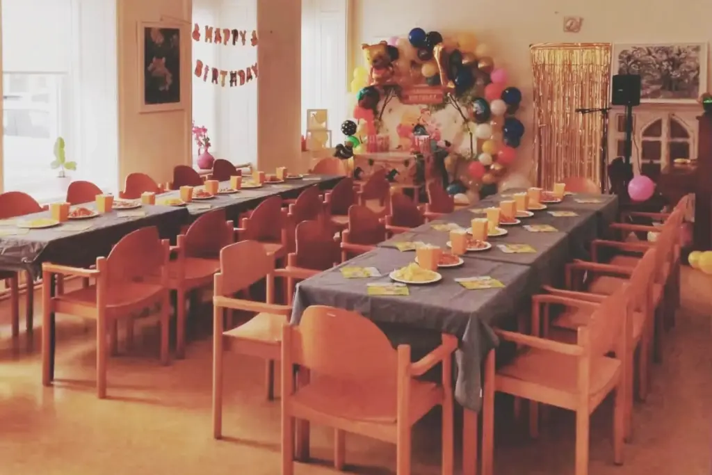 Eine Geburtstagsparty mit langen Tischen mit blauen Tischdecken, umgeben von Holzstühlen, jeder Platz mit orangefarbenen Tassen und Tellern ausgestattet. Im Hintergrund hängen Luftballondekorationen und ein Happy-Birthday-Banner.