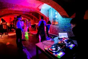 Ein DJ spielt Musik auf einem Mischpult, während die Leute in einem schwach beleuchteten, gemauerten Raum mit bunten Lichtern und einer lebhaften Partyatmosphäre tanzen und sich unterhalten.