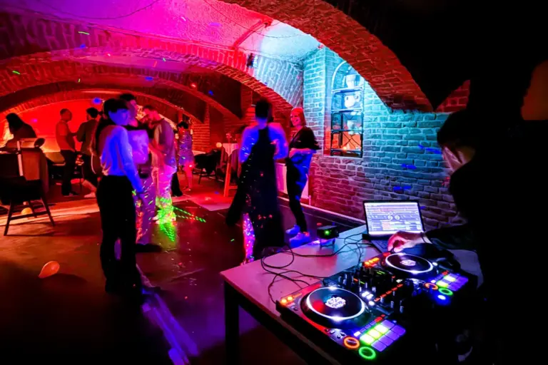 Ein DJ spielt Musik auf einem Mischpult, während die Leute in einem schwach beleuchteten, gemauerten Raum mit bunten Lichtern und einer lebhaften Partyatmosphäre tanzen und sich unterhalten.
