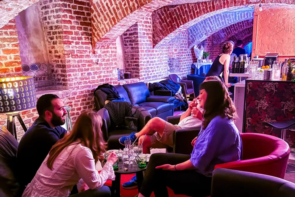 Vier Personen sitzen um einen Tisch mit Getränken in einer gemütlichen, gemauerten Lounge und unterhalten sich. Eine weitere Person steht an der Bar im Hintergrund. Die Beleuchtung ist warm und farbenfroh und schafft eine entspannte Atmosphäre.