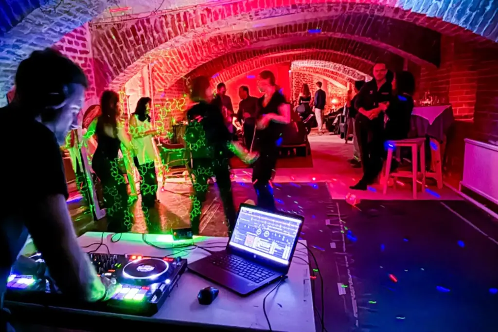 Ein DJ spielt Musik mit einem Controller in einem Gewölbe aus Ziegelsteinen, in dem bunte Lichter leuchten, während die Leute tanzen und sich im Hintergrund auf einer lebhaften Party unterhalten.