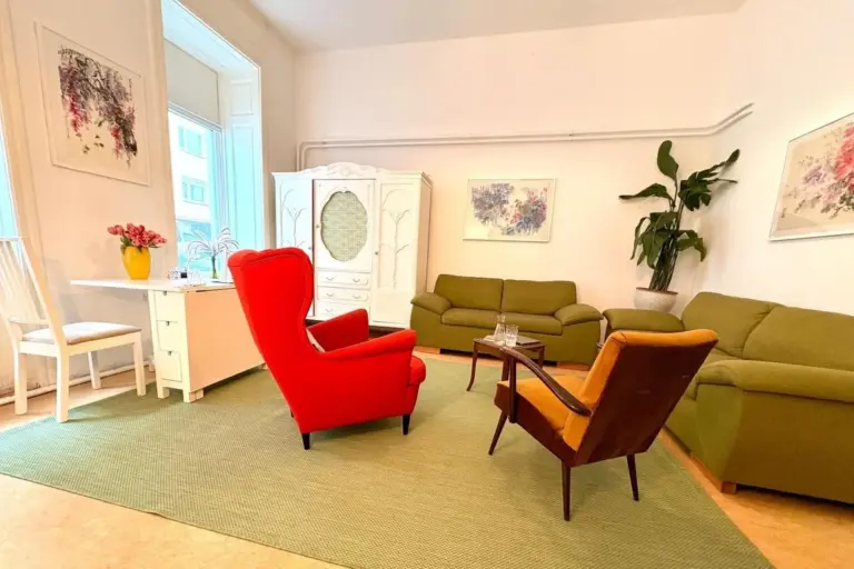 Helles Wohnzimmer mit grünen Sofas, einem kräftigen roten Sessel, einem braunen Stuhl, einem weißen Schreibtisch und Kleiderschrank, grünem Teppich, großer Topfpflanze und bunten Kunstwerken an den hellen Wänden. Sonnenlicht strömt durch ein hohes Fenster.