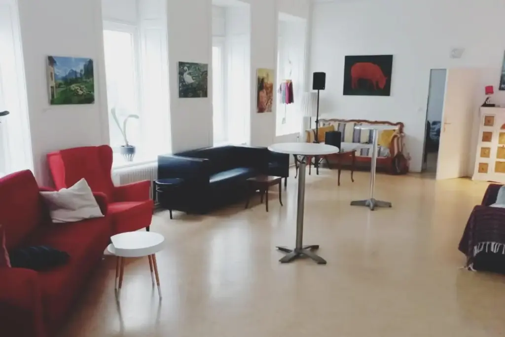 Ein heller, geräumiger Raum mit großen Fenstern, einem roten Sessel, einem schwarzen Sofa, runden Tischen mit weißen Platten und verschiedenen Kunstwerken an weißen Wänden. Der Boden ist hell, und am Fenster steht eine Topfpflanze.