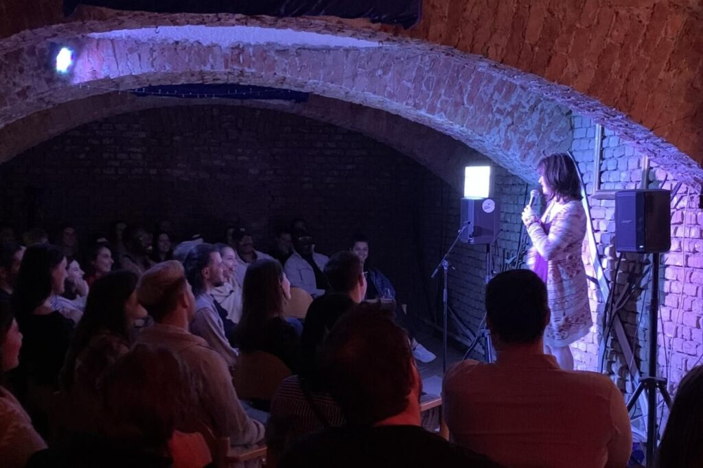 Ein Comedian tritt auf der Bühne mit einem Mikrofon in einem schwach beleuchteten Raum mit Ziegelwänden auf - perfekt für alle, die den Partykeller mieten Wien besuchen wollen - und sieht sich einem Publikum gegenüber, das dicht beieinander sitzt und aufmerksam zusieht.