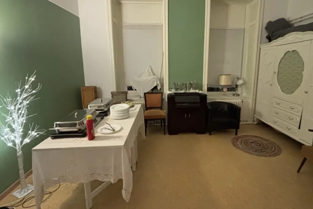 In einem Raum mit grünen und weißen Wänden steht ein Tisch mit Chafing Dishes, Tellern und Utensilien. In der Nähe steht ein weißer Zierbaum, und an den Wänden stehen verschiedene Möbel, darunter Stühle, Schränke und ein Kleiderschrank.