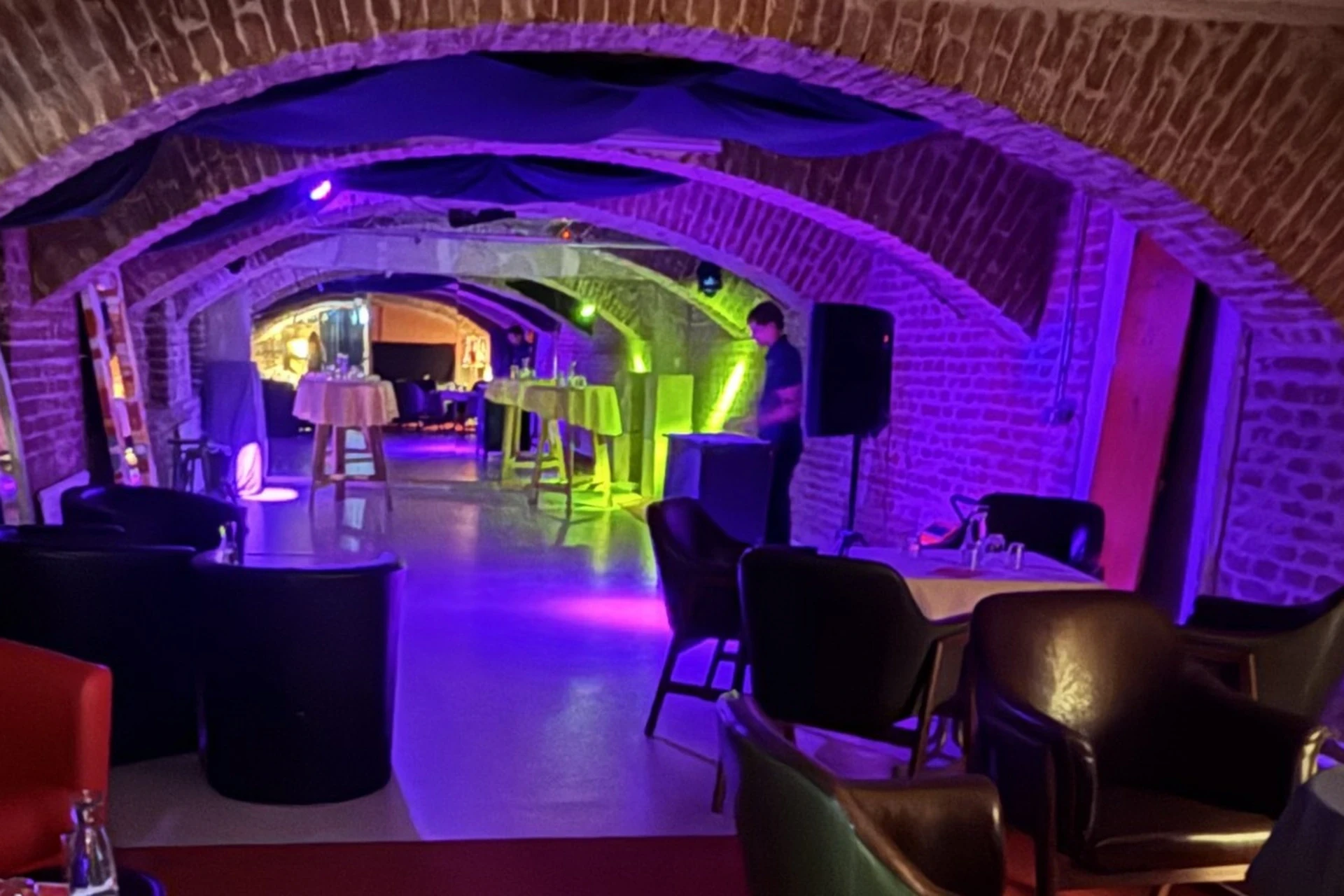 Eine schwach beleuchtete Lounge mit gewölbten Ziegeldecken, modernen Tischen und Stühlen und violetter Umgebungsbeleuchtung. Eine Person steht neben einem Lautsprecher, und im Hintergrund sind mehrere Tische aufgestellt.