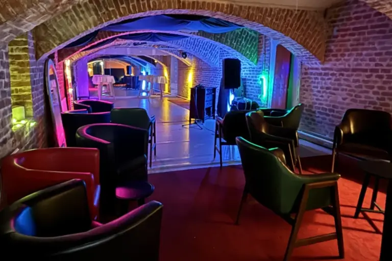dasWallenstein Eine stilvolle Lounge mit Gewölbedecken aus Ziegeln, farbenfroher Beleuchtung, schwarzen Stühlen und kleinen Tischen, die eine gemütliche und lebendige Atmosphäre in einer unterirdischen Umgebung schaffen.