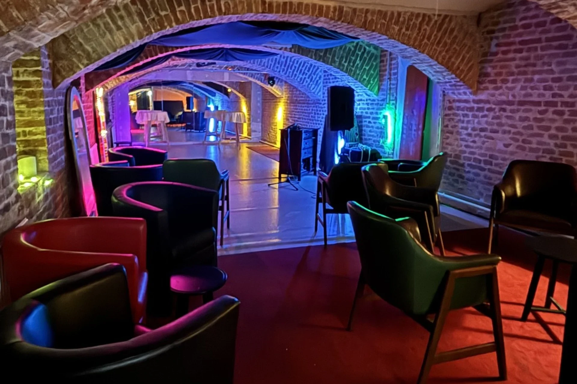 dasWallenstein Eine stilvolle Lounge mit Gewölbedecken aus Ziegeln, farbenfroher Beleuchtung, schwarzen Stühlen und kleinen Tischen, die eine gemütliche und lebendige Atmosphäre in einer unterirdischen Umgebung schaffen.