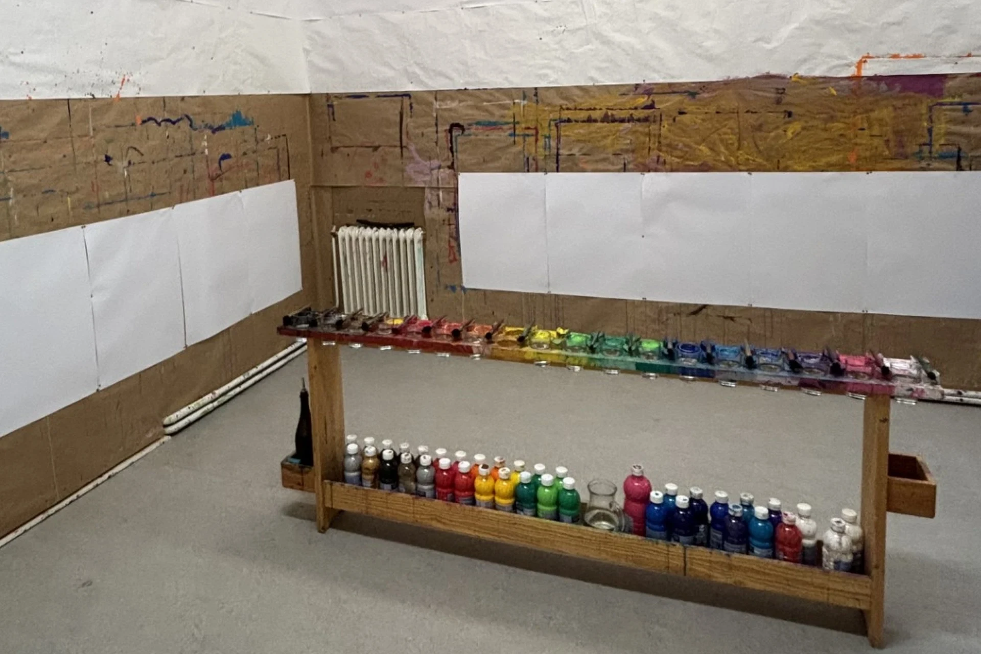 Auf einem Holztisch stehen bunte Farbtöpfe und Pinsel, die nach Farben geordnet sind. Leere weiße Papiere sind mit Klebeband an die braunen, papierbedeckten Wände geklebt, was auf ein Kunstatelier schließen lässt, das für eine Malaktion bereit ist.