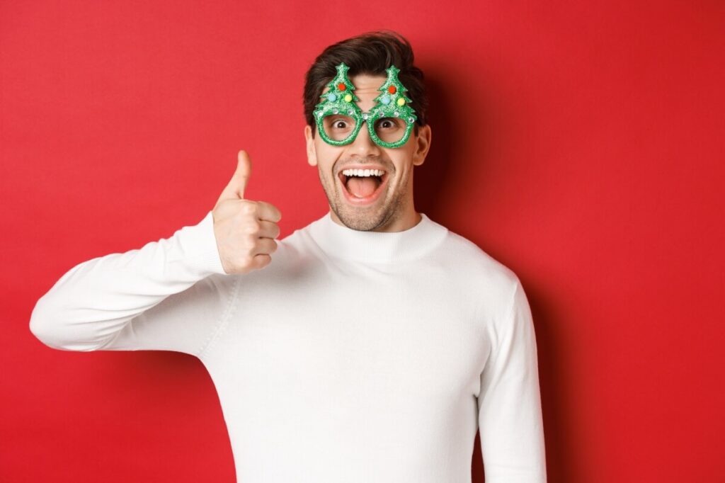 Ein lächelnder Mann mit einer neuartigen Weihnachtsbaumbrille und einem weißen Pullover steht vor einem roten Hintergrund und zeigt den Daumen nach oben - die perfekte Inspiration für Last-Minute-Geschenkideen in dieser Weihnachtszeit.