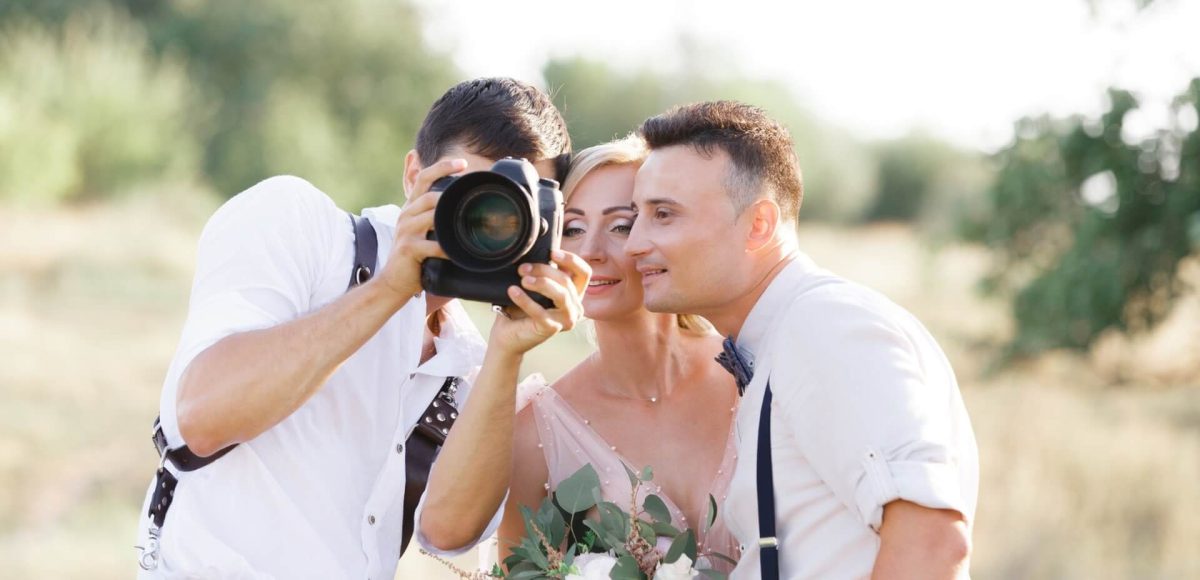 Ein Fotograf für Hochzeit zeigt Braut und Bräutigam ihr Foto mit seiner Kamera. Das lächelnde Paar, das einen Blumenstrauß in der Hand hält, steht eng beieinander im Freien in schönem natürlichem Licht.
