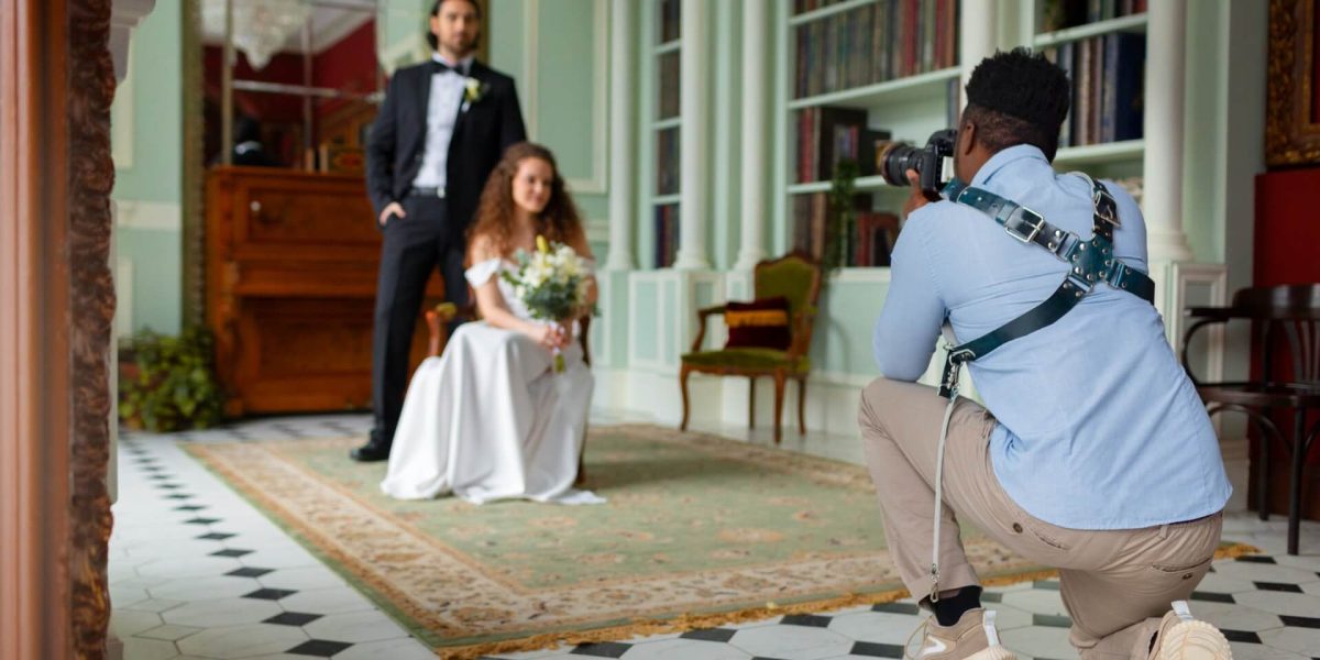Ein Fotograf für Hochzeit kniet nieder und fotografiert eine Braut mit ihrem Brautstrauß, während der Bräutigam hinter ihr in einem eleganten, mit hohen Bücherregalen und gemusterten Böden geschmückten Raum im Vintage-Stil steht.